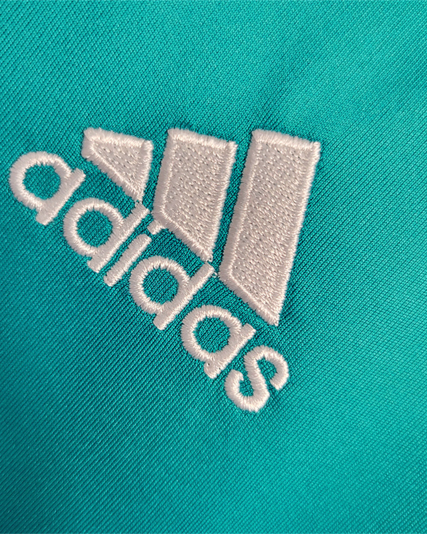 Dettaglio ravvicinato dello stemma del Adidas ricamato in bianco su tessuto turchese.