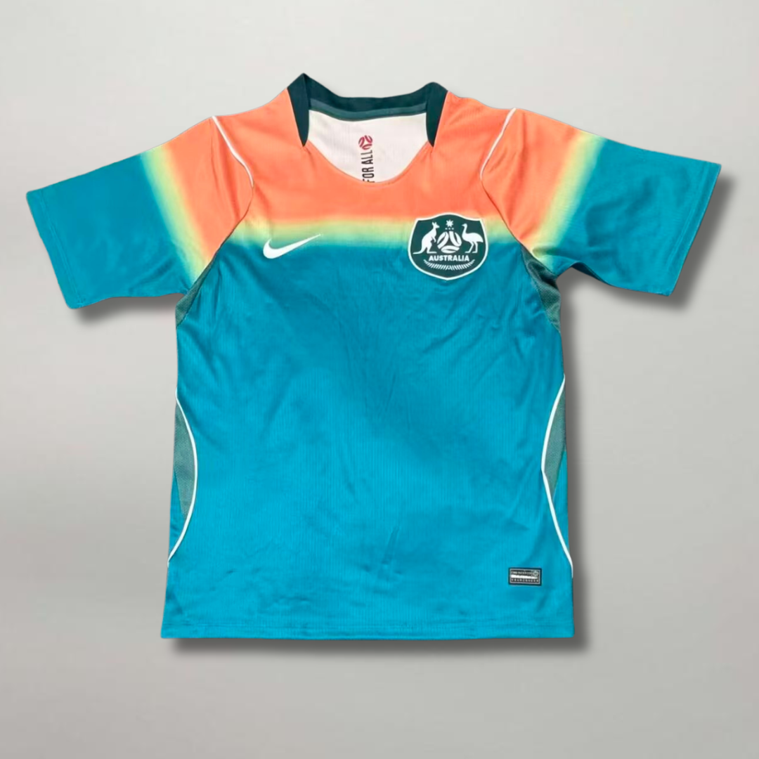 Fronte della maglia away dell'Australia 2026 turchese con spalle sfumate oro e arancione e stemma sul petto