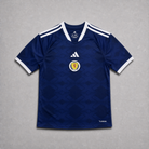 Maglia home Scozia Coppa del Mondo 2026 — vista frontale su superficie grigia, tessuto blu navy con colletto a V con bordino bianco, stemma della federazione scozzese sul petto sinistro, maniche con profili bianchi
